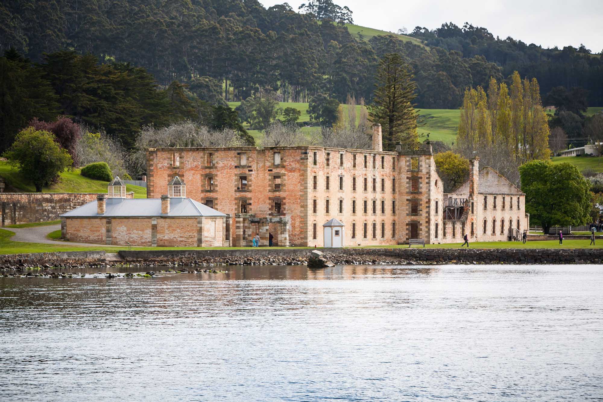 A historic brick penitentiary complex situated on the waterfront at Port Arthur, Tasmania, featuring multiple stone and brick buildings with characteristic 19th-century colonial architecture set against forested hills and calm waters.