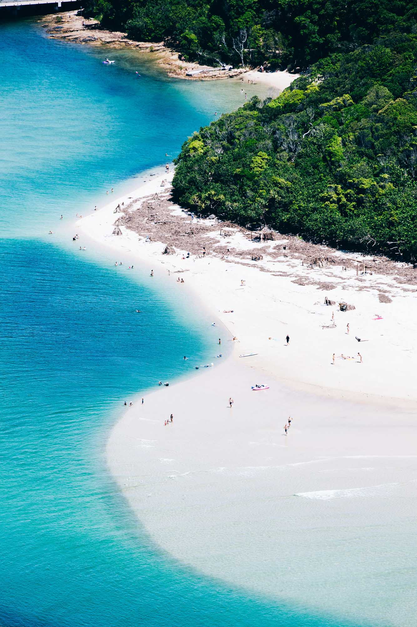 An aerial view of a stunning curved white sand beach with crystal-clear turquoise waters, featuring small groups of beachgoers swimming and relaxing. Dense tropical vegetation and lush green forest backdrop the crescent-shaped shoreline, creating a picturesque tropical paradise destination.