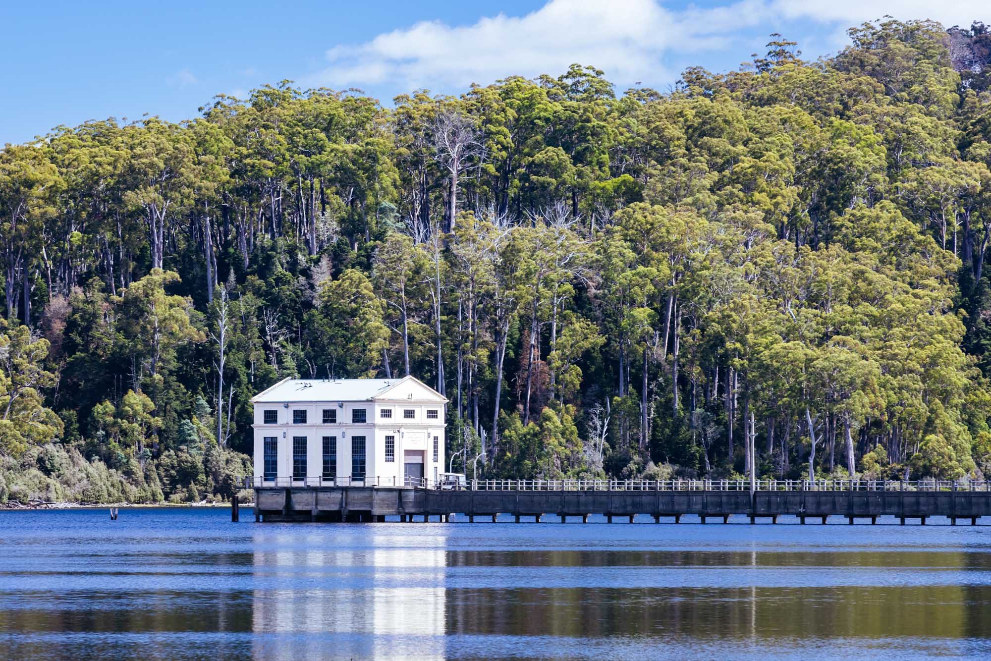 A striking white neoclassical boathouse structure stands on the shore of Lake St Clair in Tasmania, backed by a dense native eucalyptus forest. The serene waters reflect the clear blue sky, creating a picturesque scene of Australia's wilderness heritage.