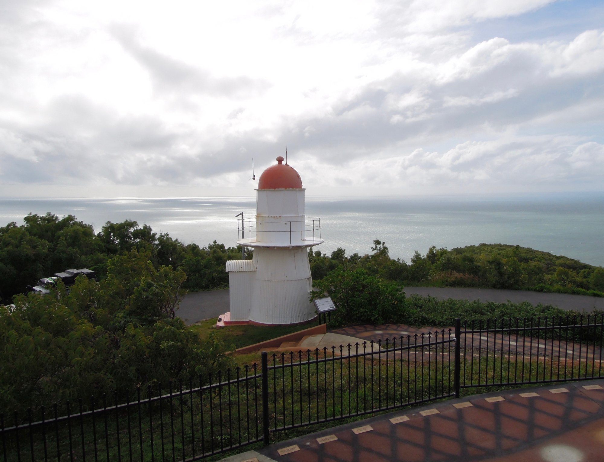 A striking white and red cylindrical lighthouse with a distinctive domed top stands on a forested headland overlooking the Coral Sea near Cooktown, Australia. The structure features navigational equipment and a protective railing, situated among lush tropical vegetation with calm waters visible in the distance.