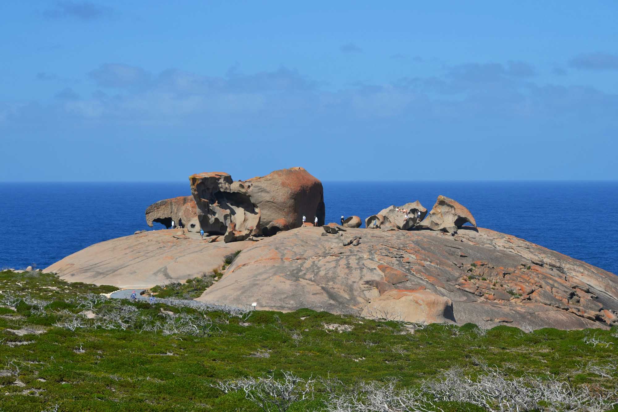 A striking granite boulder formation rises dramatically from a windswept coastal plateau overlooking the blue ocean. The massive, precariously balanced rocks are among Australia's most iconic natural landmarks, set against vibrant green vegetation and a clear sky.