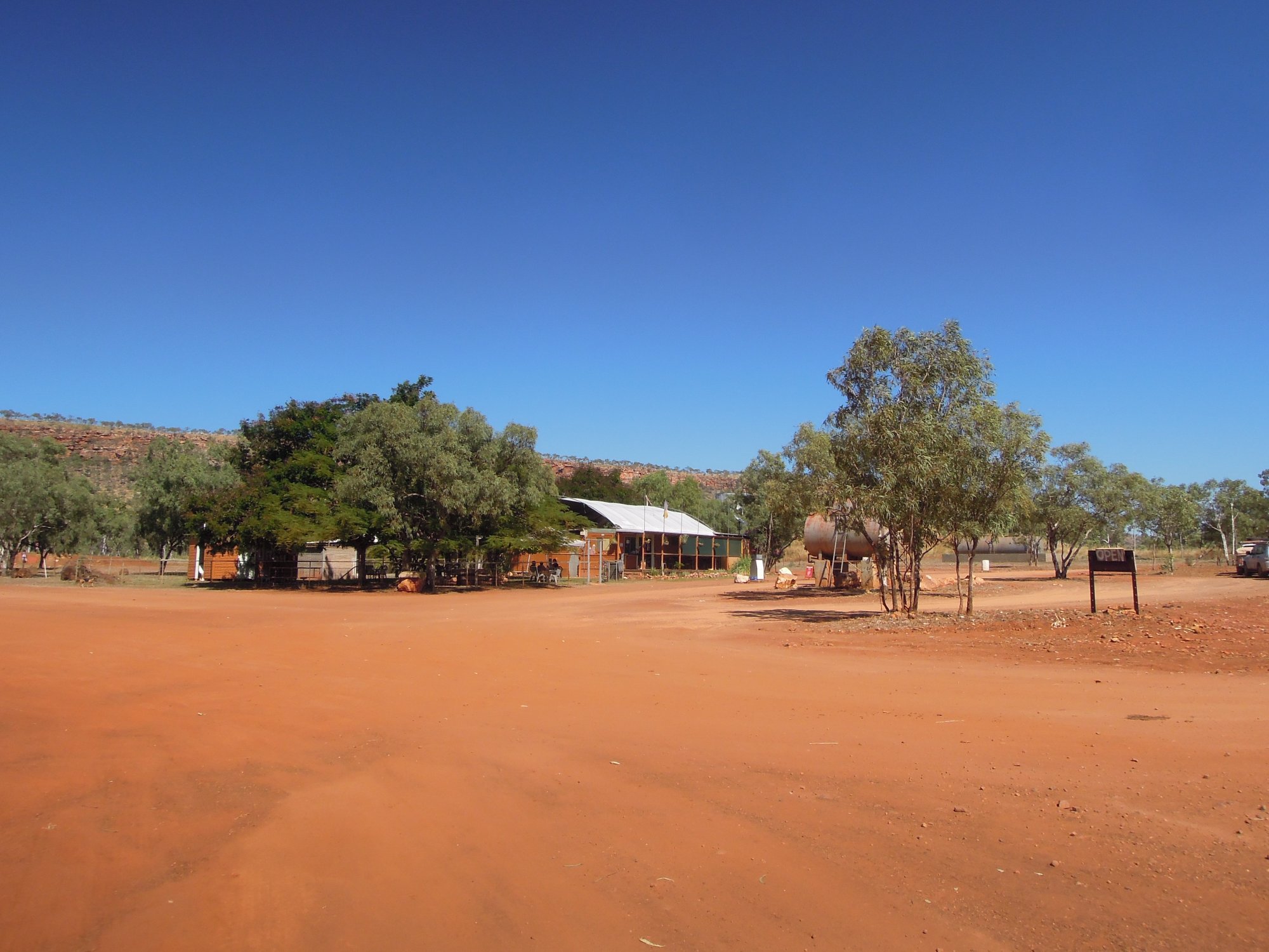 A rustic, isolated general store and rest stop located along the Gibb River Road in the remote Kimberley region of Western Australia. The weathered orange-brown corrugated iron building sits beneath sprawling eucalyptus trees on red earth, with a water tank visible, serving as a crucial waypoint for travelers crossing the outback.