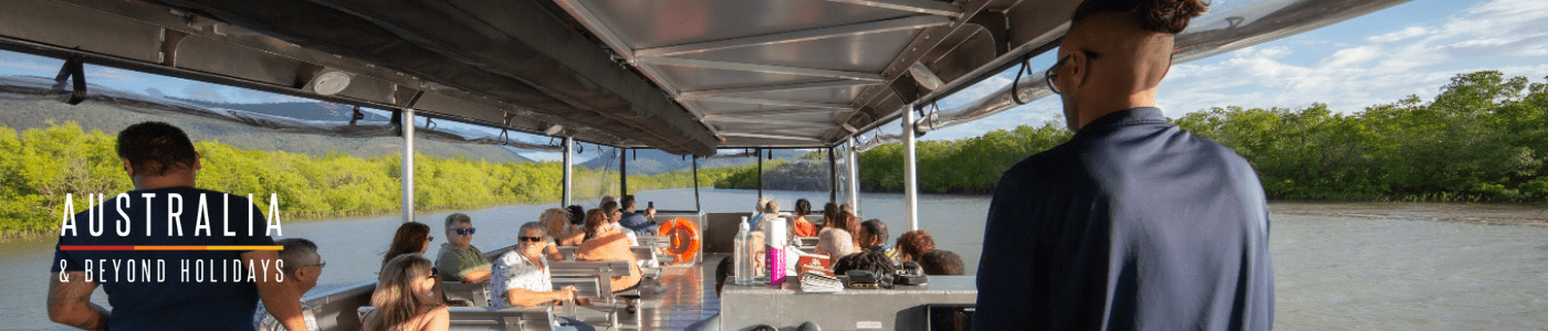 A scenic aerial view of a traditional tour boat navigating through winding mangrove waterways near Cairns, Queensland. The lush green mangrove forest is bordered by a meandering river that opens to a coastal bay with distant mountains and a charming port town visible in the background.