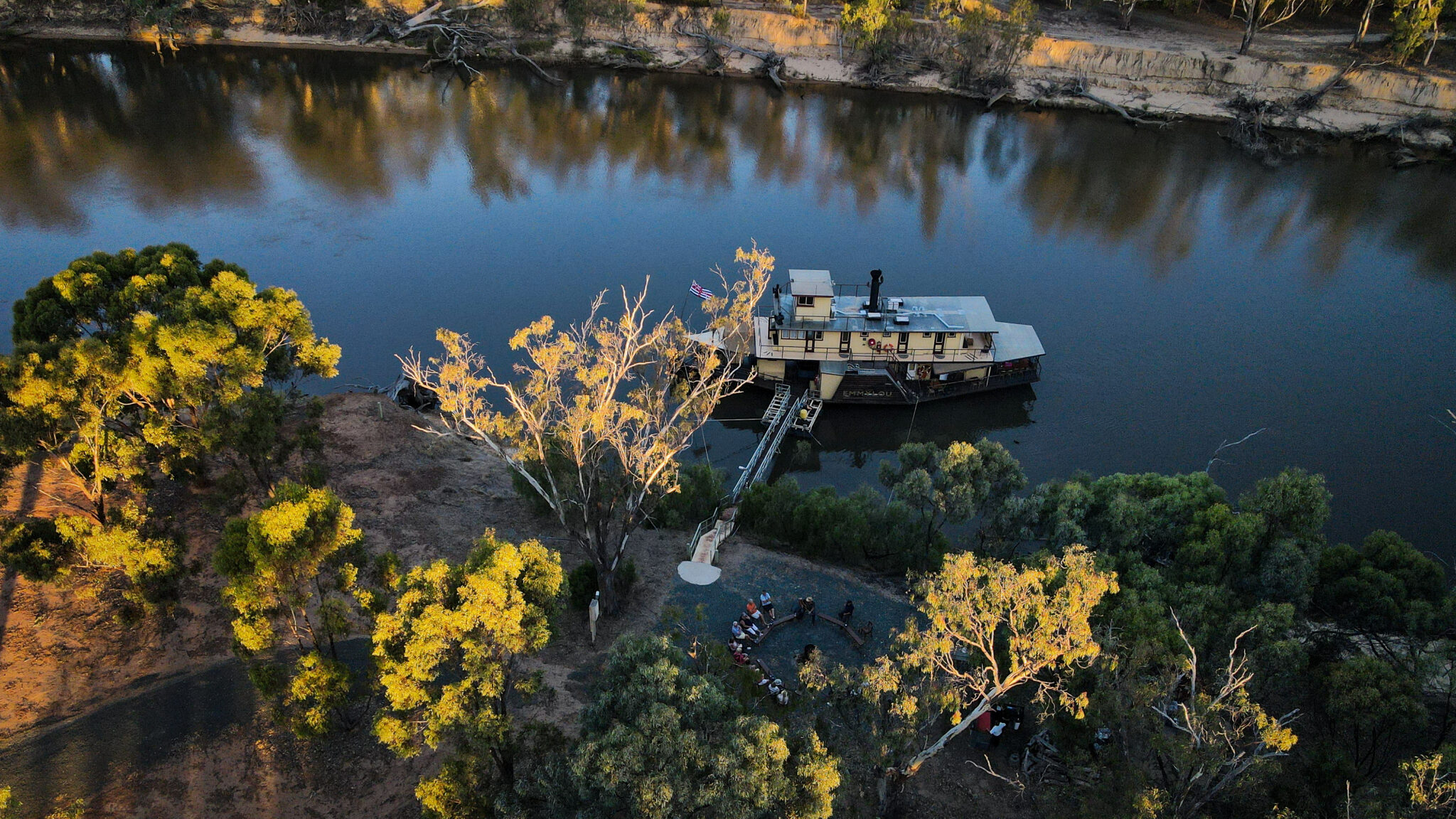 An aerial view of a multi-deck river cruise ship moored at a sandy riverbank landing with passengers gathered on shore surrounded by golden-lit eucalyptus trees. The vessel features a distinctive white and cream livery with multiple deck levels, connected to the landing via a gangway, on a calm, mirror-like river with dramatic cliff faces visible in the background.