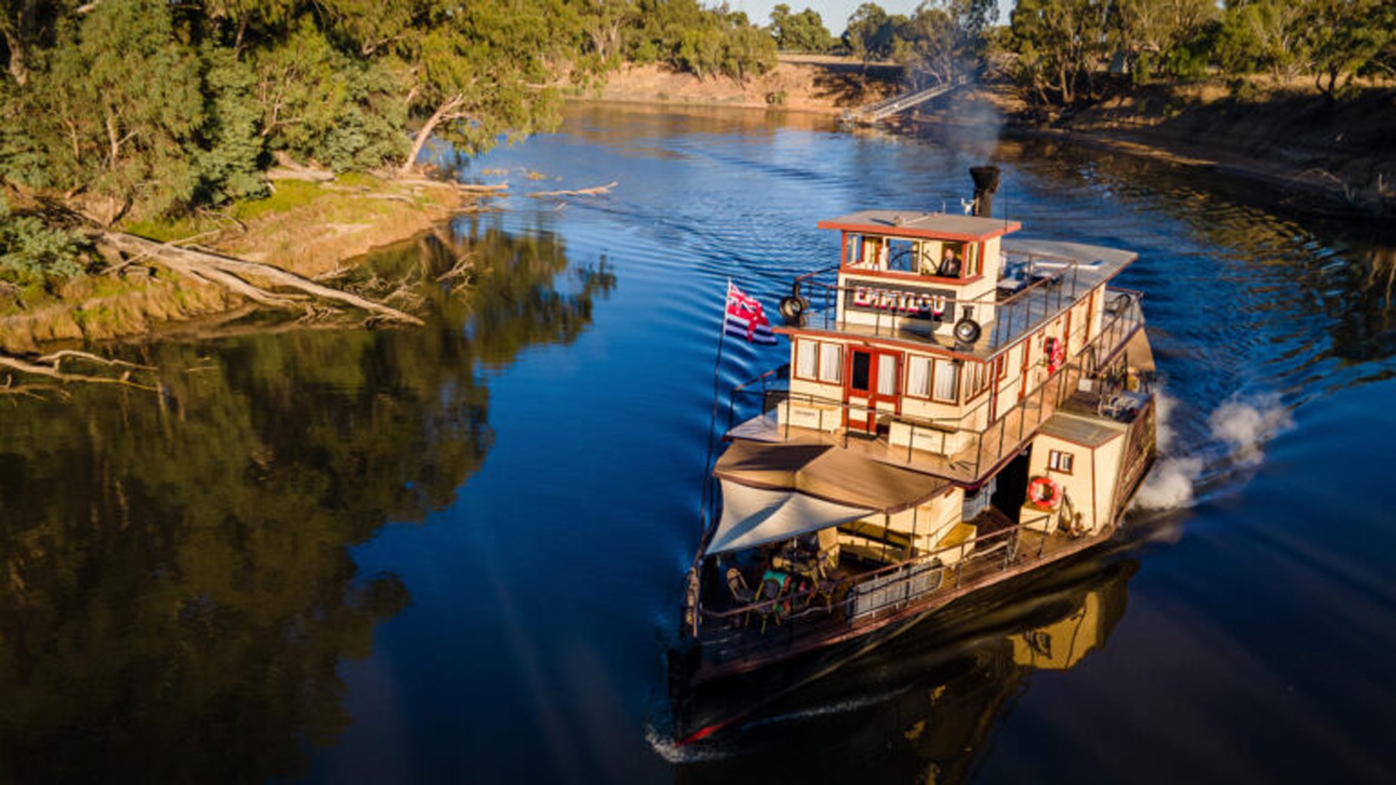 A distinctive multi-deck riverboat with cream and orange/red coloring navigates a serene river lined with native eucalyptus trees and natural bushland. The vessel features multiple outdoor decks, railings, and what appears to be an Australian flag, characteristic of river cruise operations in inland Australia.