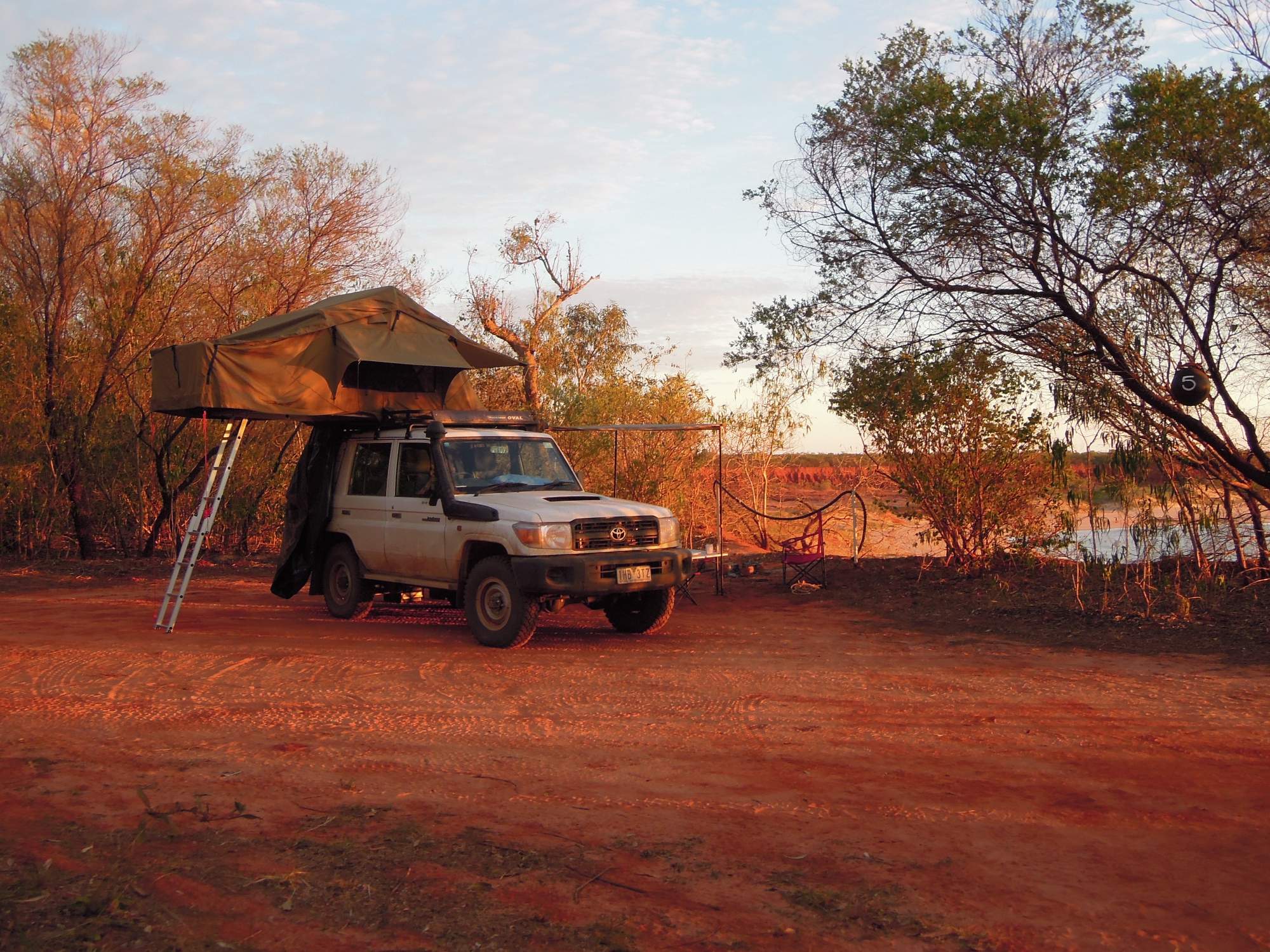 A Toyota Land Cruiser equipped with a rooftop tent is parked at a remote campsite in the Australian outback, surrounded by sparse vegetation and red earth. The scene captures adventure travel and off-road camping in an arid, expansive landscape during golden hour.