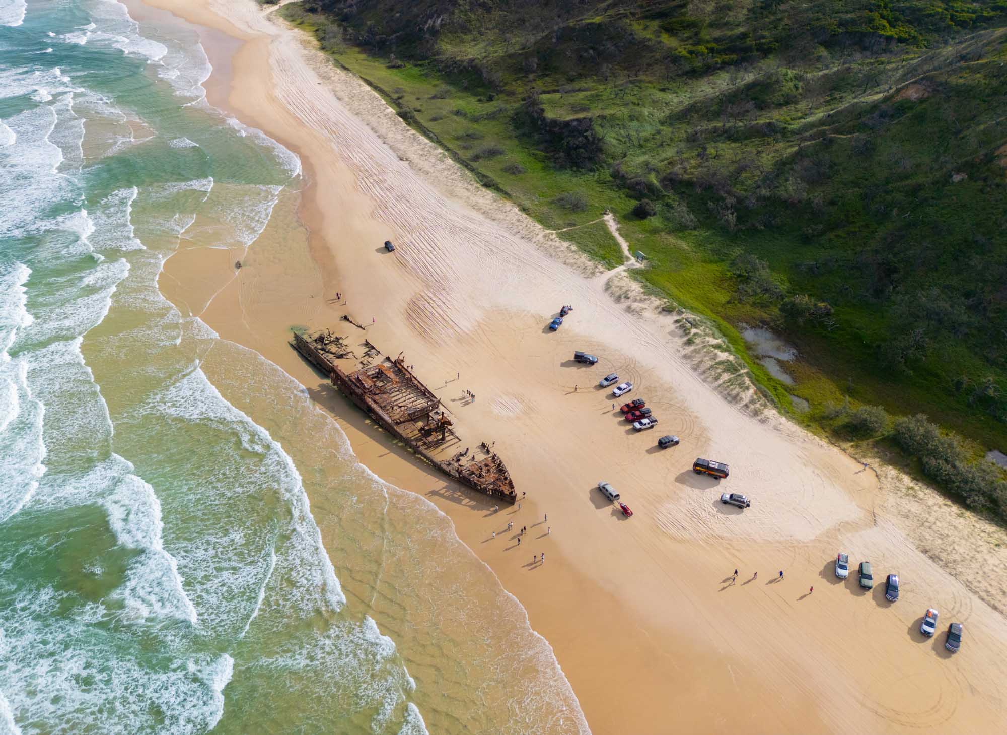 An aerial view of the abandoned S.S. Maheno shipwreck resting on the sandy beach of K'gari (Fraser Island) in Queensland, Australia, surrounded by parked vehicles and with waves washing ashore and lush green vegetation visible inland.