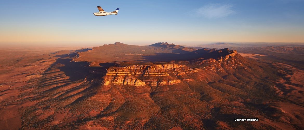 An aerial photograph taken from a small aircraft flying over Wilpena Pound, showcasing the dramatic mountain ranges and rugged terrain bathed in golden morning light. The landscape displays the characteristic ridged formations and vast arid plains typical of South Australia's Flinders Ranges.