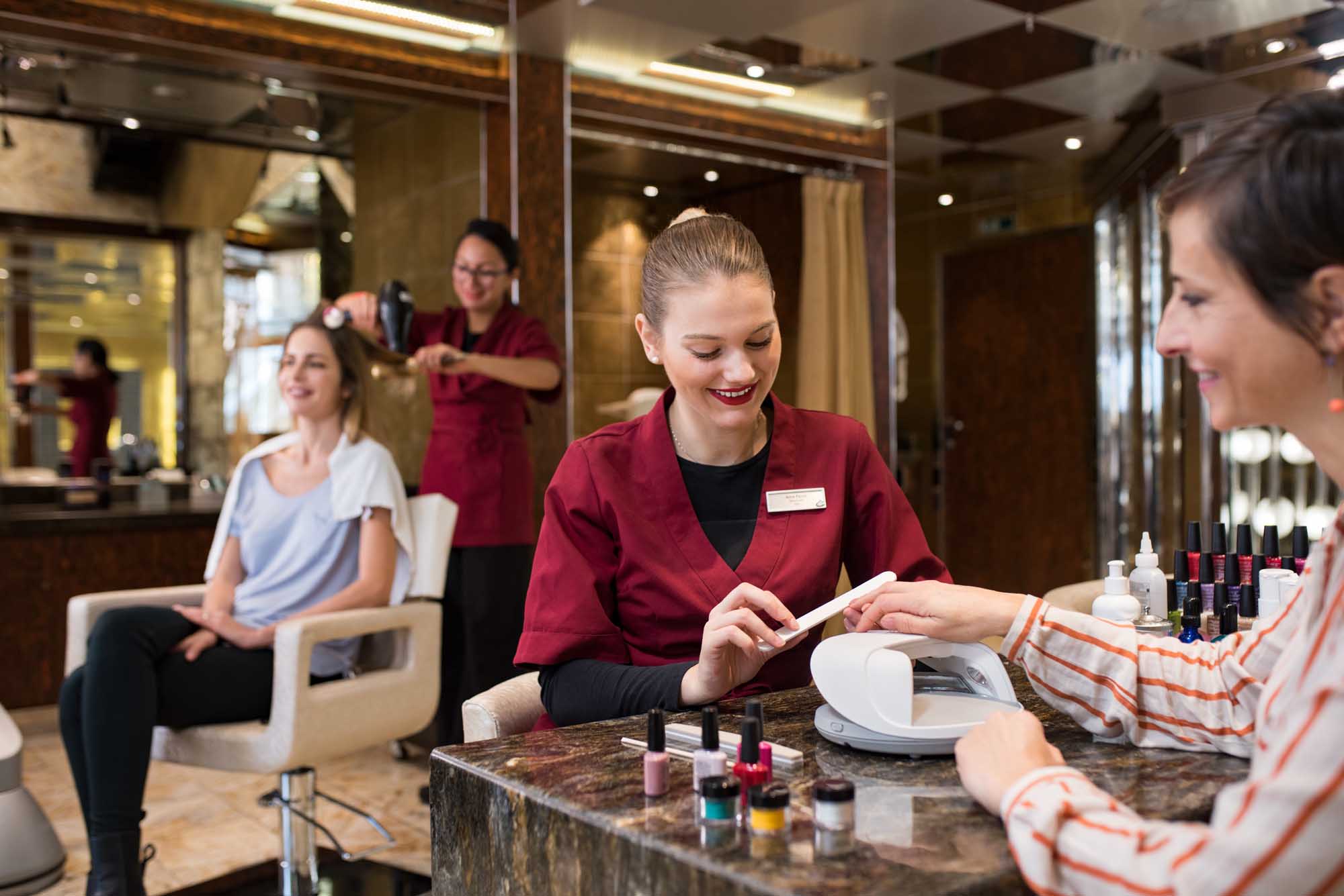 A professional spa receptionist in a burgundy uniform processes a booking or payment for a guest at an elegant salon counter, with nail polish displays and beauty products visible. The luxurious interior features mirrored walls, warm lighting, and multiple staff members attending to clients in the background, creating an upscale cruise ship spa environment.
