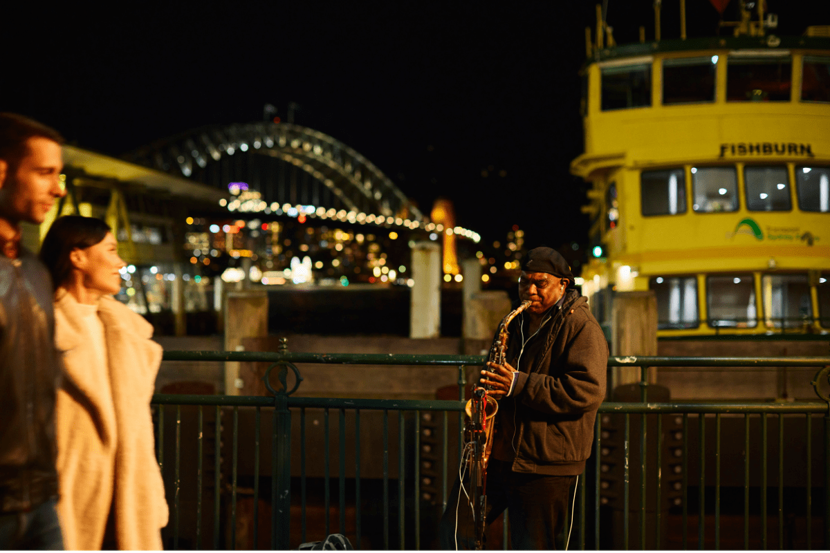 A saxophone player performs on a waterfront promenade at night, with the illuminated Sydney Harbour Bridge visible in the background and a yellow Fishburn ferry visible to the right. Pedestrians stroll past the street musician in this iconic Sydney harbour scene.