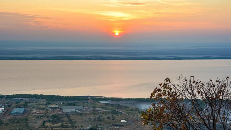 A breathtaking golden-hour sunset view over the Kimberley region's coastline and inland landscape, with warm orange and amber tones reflecting across calm waters and revealing scattered settlements and agricultural areas below.