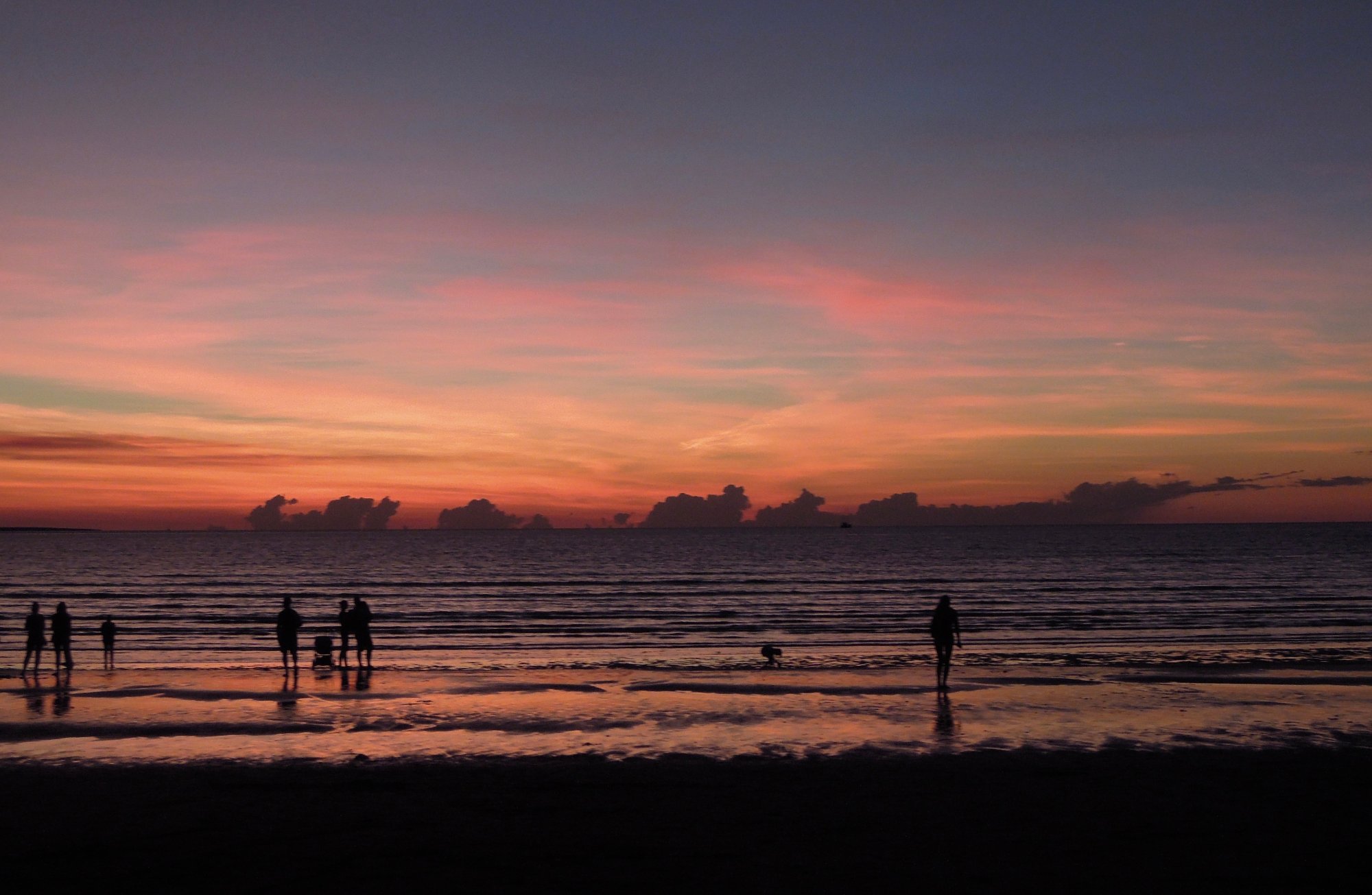A stunning tropical sunset over the Indian Ocean at Mindil Beach in Darwin, Australia, with silhouetted beachgoers wading in the shallow waters as vibrant orange and pink hues paint the sky and reflect off the wet sand.
