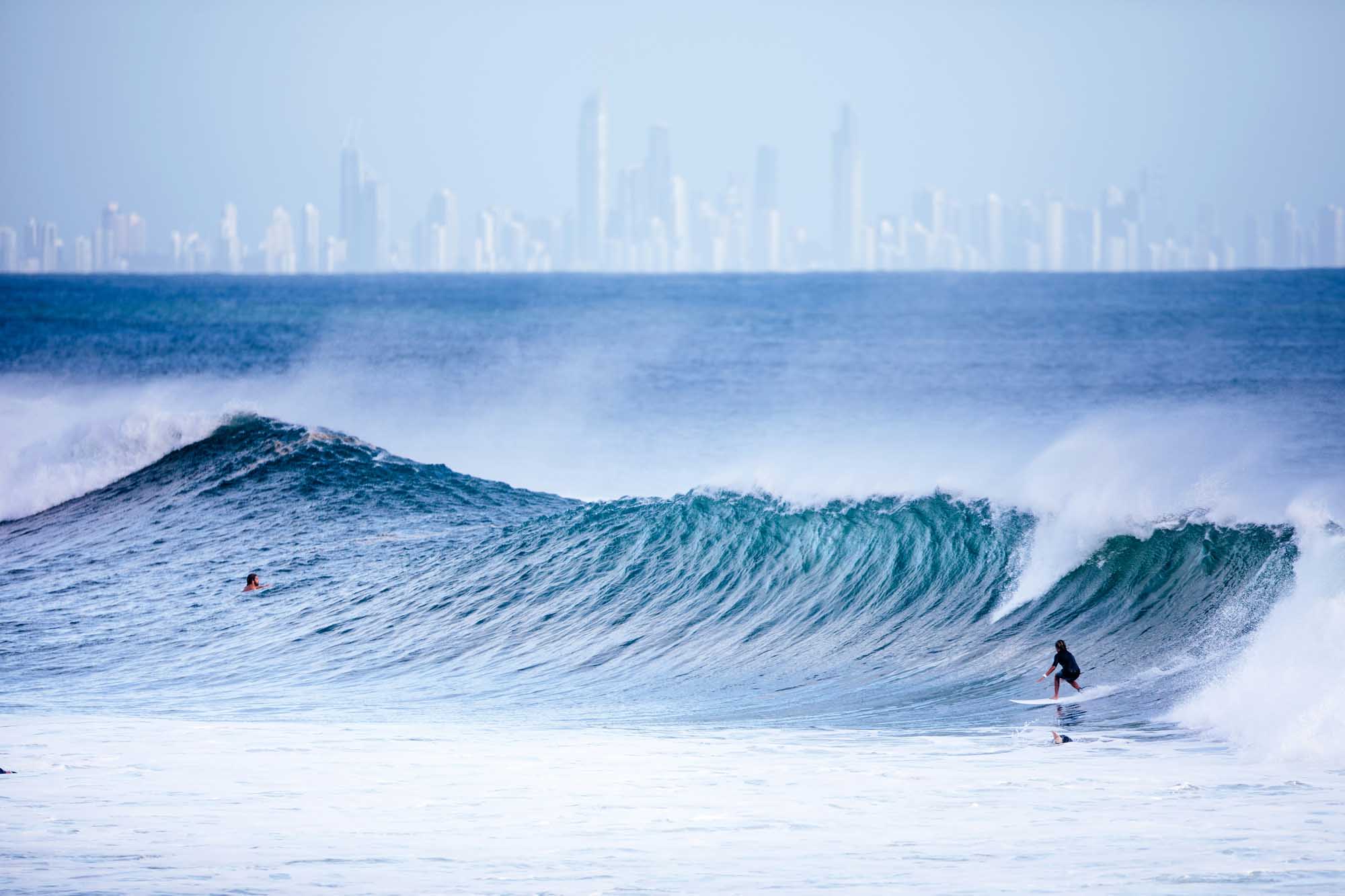 A dynamic surfing scene featuring multiple surfers riding large ocean waves with the iconic Gold Coast city skyline visible in the misty background across the water. The image captures the contrast between the natural ocean swells and urban development.