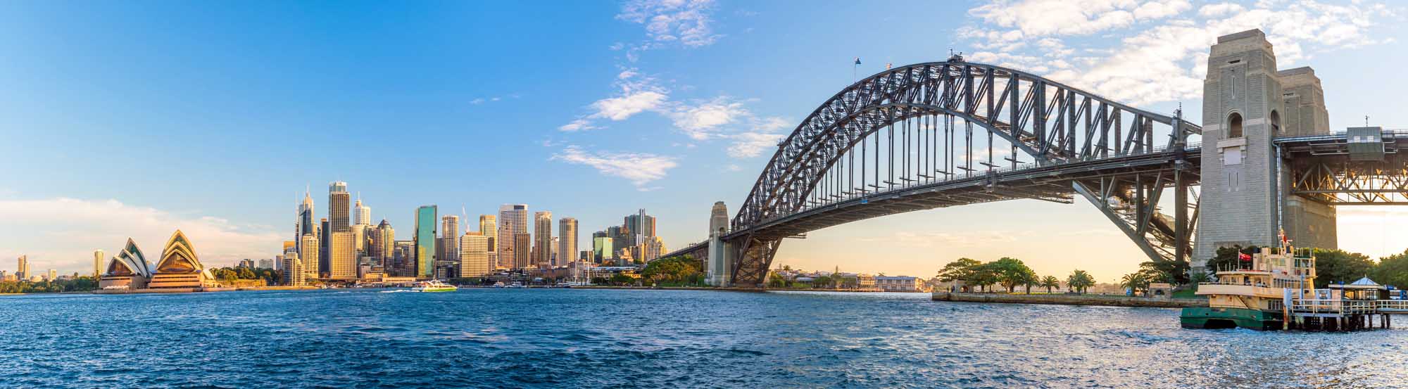 A panoramic view of Sydney, Australia's iconic skyline featuring the Sydney Opera House, downtown skyscrapers, and the historic Sydney Harbour Bridge spanning across blue harbour waters under a partly cloudy sky.