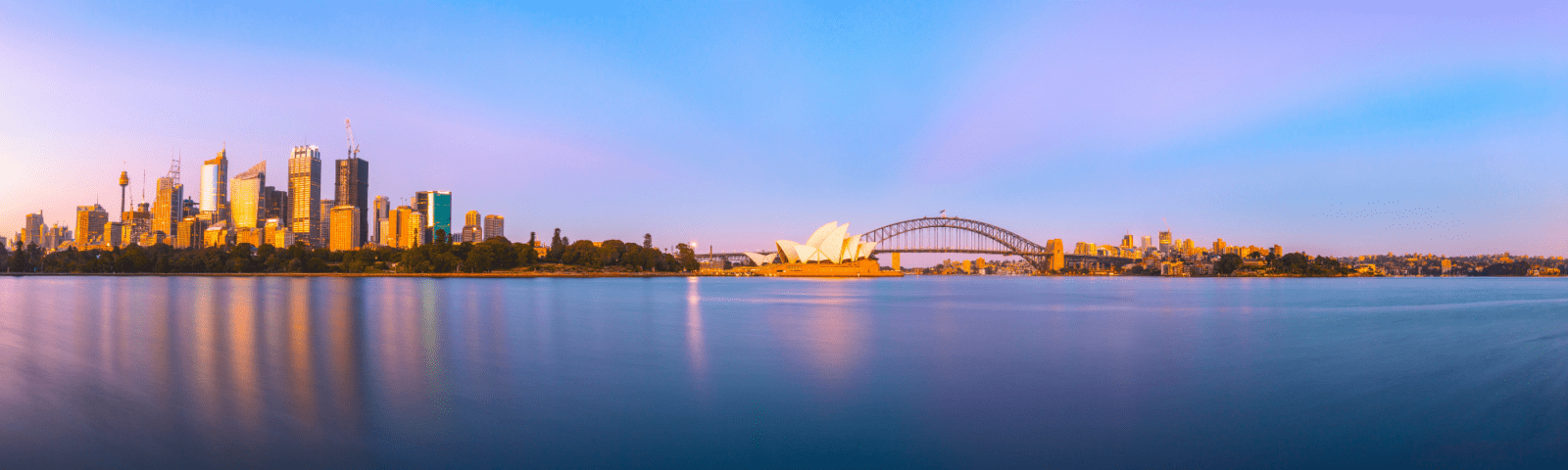 A stunning panoramic view of Sydney's iconic skyline at dawn, featuring the illuminated Central Business District towers, the distinctive Sydney Opera House, and the Sydney Harbour Bridge reflected in the calm blue waters. The image captures the golden light of early morning with a serene waterfront atmosphere.