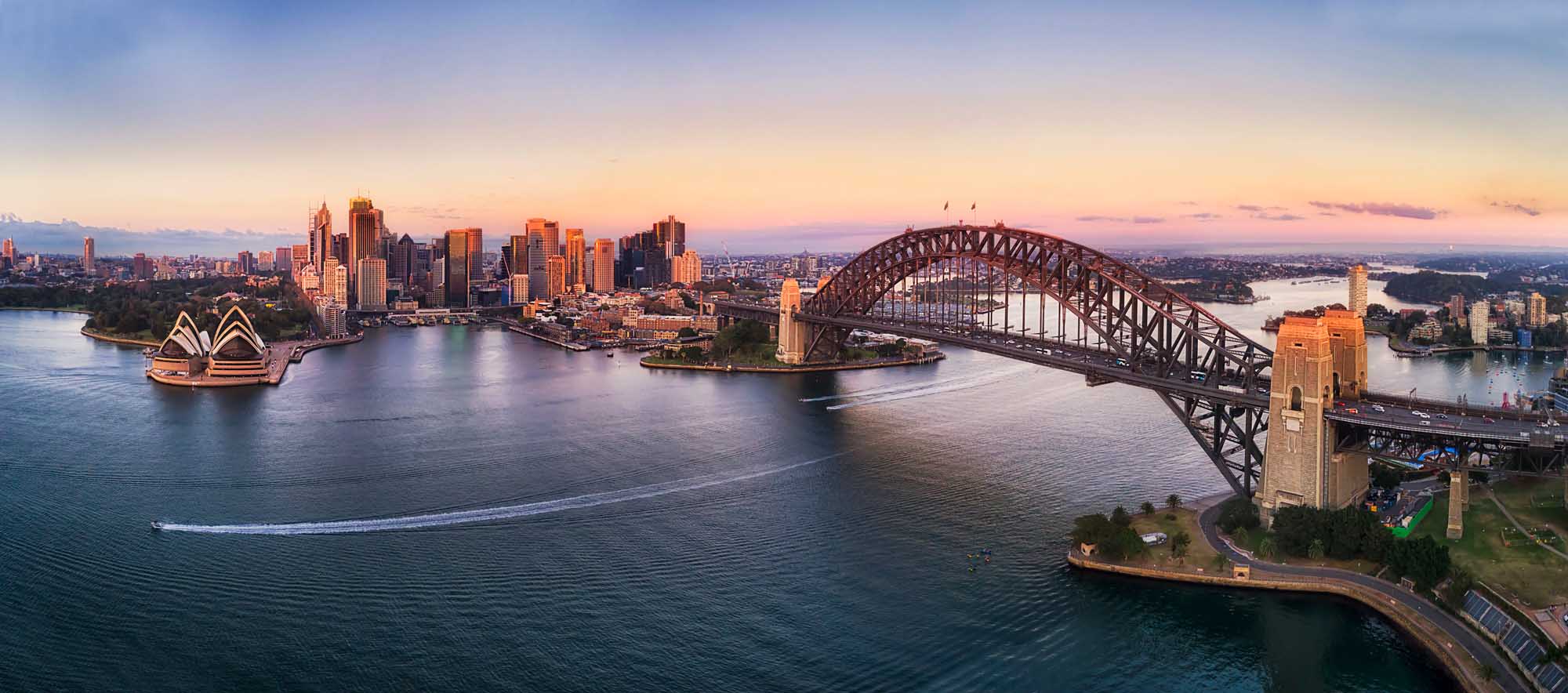 An aerial panoramic view of Sydney's iconic waterfront at dawn, featuring the illuminated Sydney Opera House on the left, the Sydney Harbour Bridge arching across the center, and the city's gleaming CBD skyscrapers bathed in warm golden light. The calm waters of Sydney Harbour reflect the soft colors of the sunrise sky, with residential areas visible beyond.