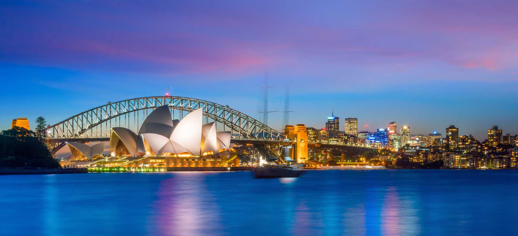 An iconic twilight view of Sydney Harbour featuring the illuminated Sydney Opera House and the Sydney Harbour Bridge, with the city skyline glowing in the background and calm blue water reflecting the lights during the magical purple and blue hour.