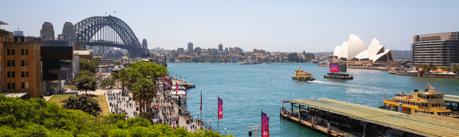 A stunning daytime view of Sydney Harbour featuring the iconic Opera House, Sydney Harbour Bridge, and vibrant turquoise waters with ferries and cruise vessels. The image captures the bustling waterfront promenade with crowds of visitors, historic colonial architecture, and modern skyscrapers lining the harbour.