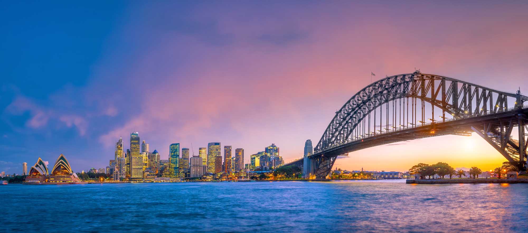 A stunning twilight view of Sydney's iconic skyline featuring illuminated skyscrapers, the distinctive Sydney Opera House on the left, and the Sydney Harbour Bridge spanning across the harbor. The scene captures the vibrant cityscape at dusk with warm golden and purple hues reflecting off the calm harbor waters.