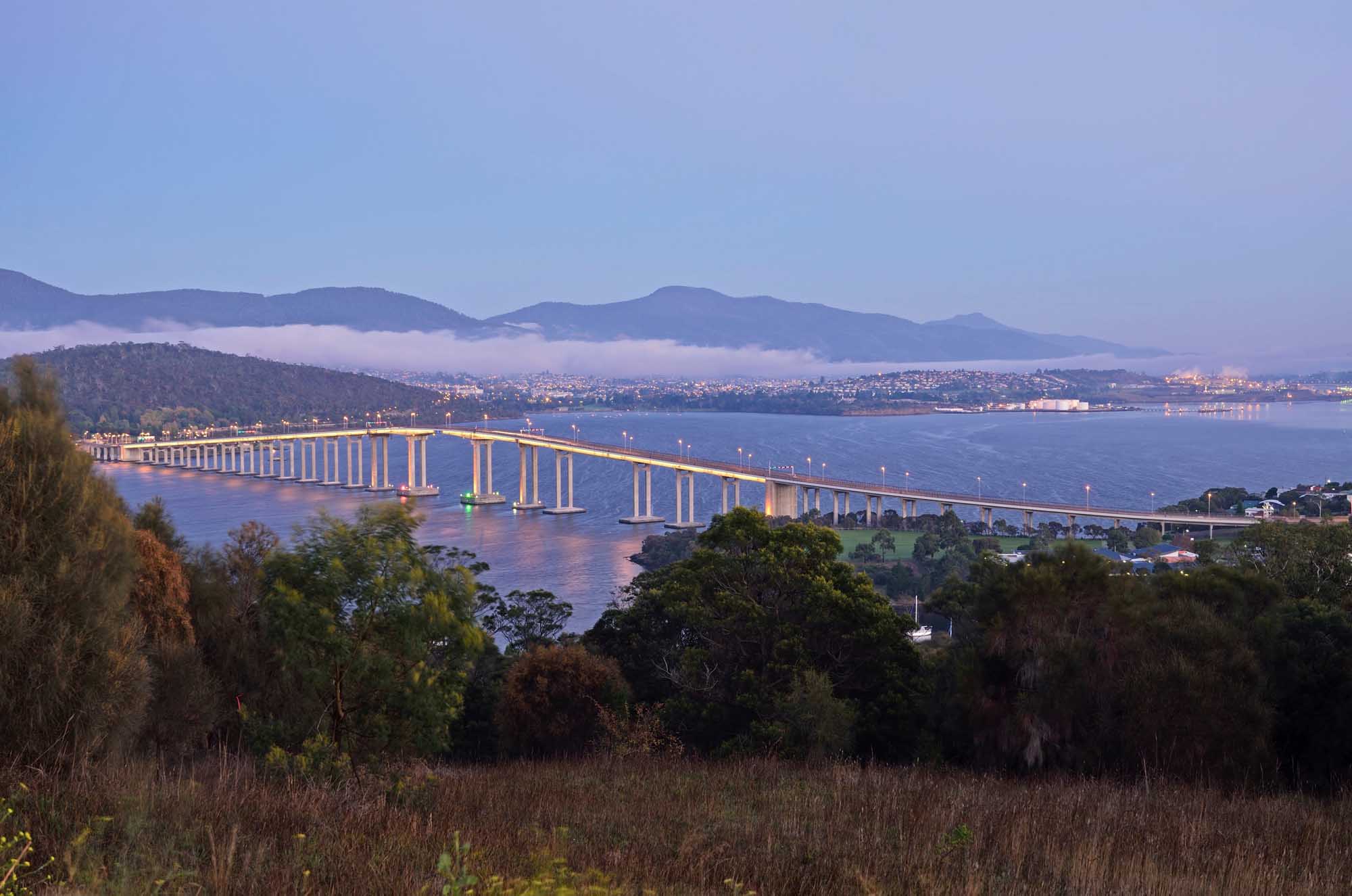The iconic Tasman Bridge illuminated at dusk spans the River Derwent in Hobart, Tasmania, with the city skyline visible across the water and mountains shrouded in mist in the background. Forested slopes frame the scene in the foreground, creating a picturesque view of this major Australian infrastructure landmark.