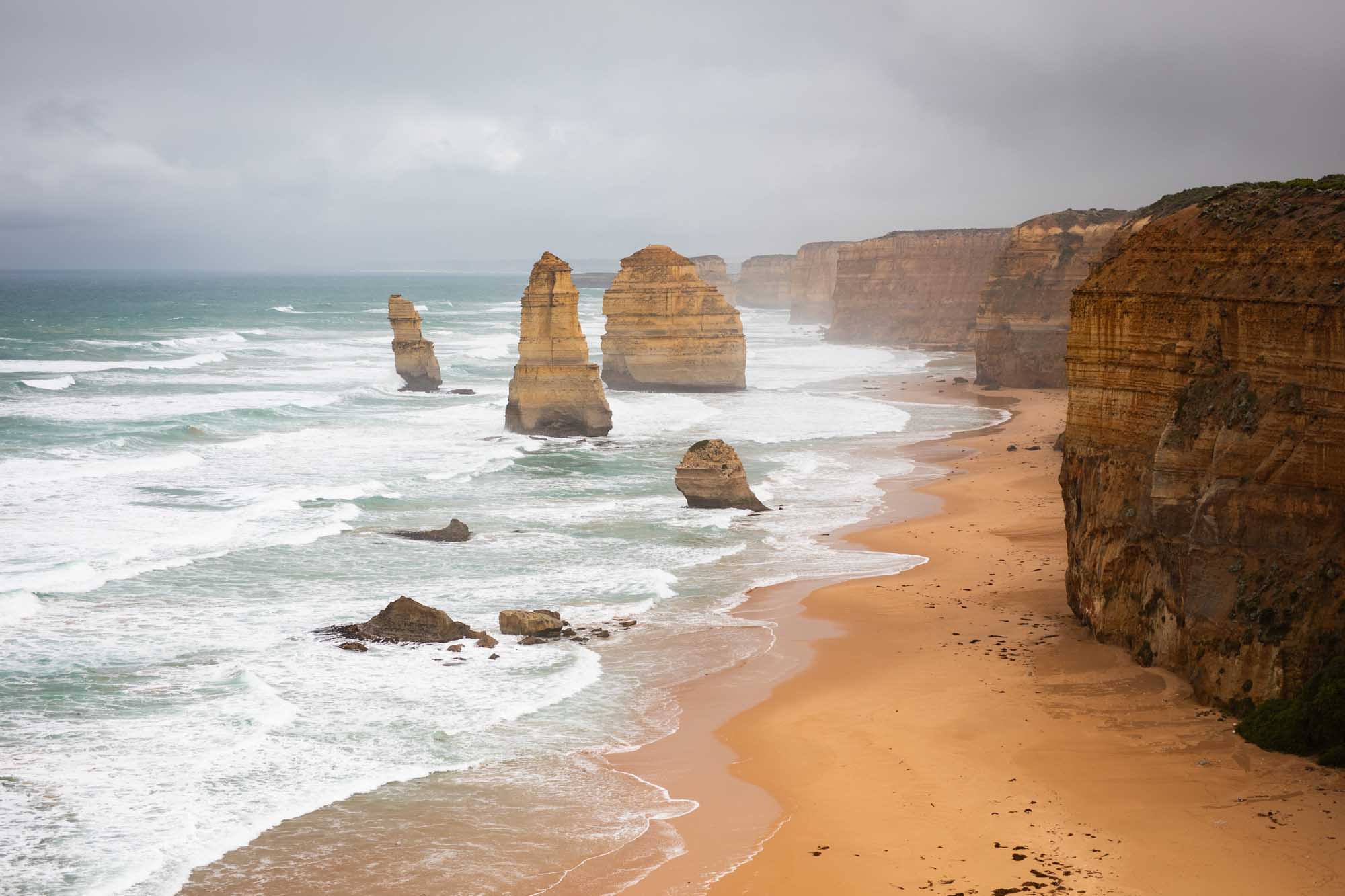 Dramatic limestone sea stacks rise majestically from a golden sandy beach along a rugged coastline, with white-capped ocean waves crashing against the shore and towering cliffs framing the composition under an overcast sky.
