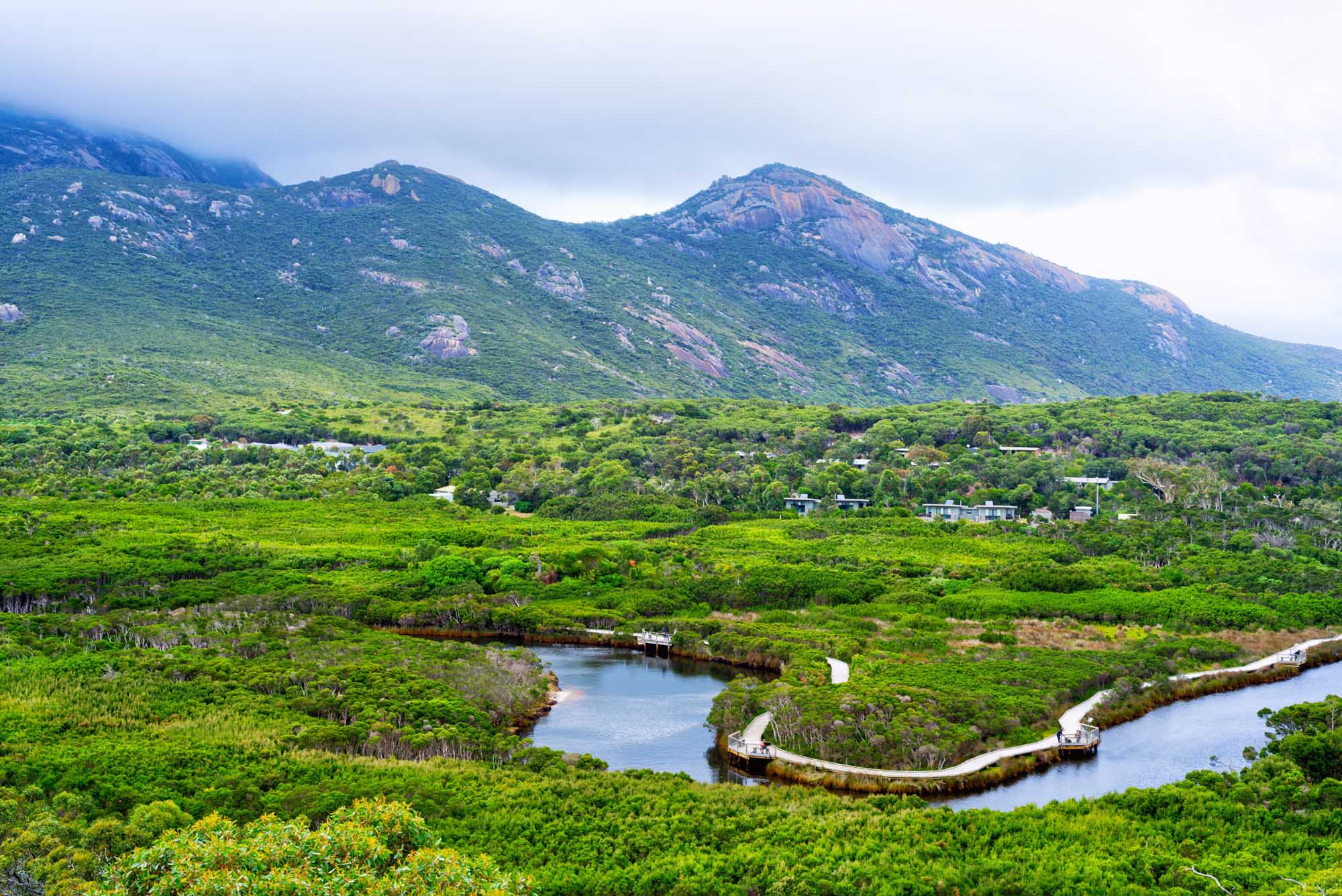 An aerial view of a scenic tidal river winding through lush green bushland at Wilsons Promontory National Park in Victoria, Australia, with distinctive granite peaks rising majestically in the background and scattered residences nestled among native vegetation.