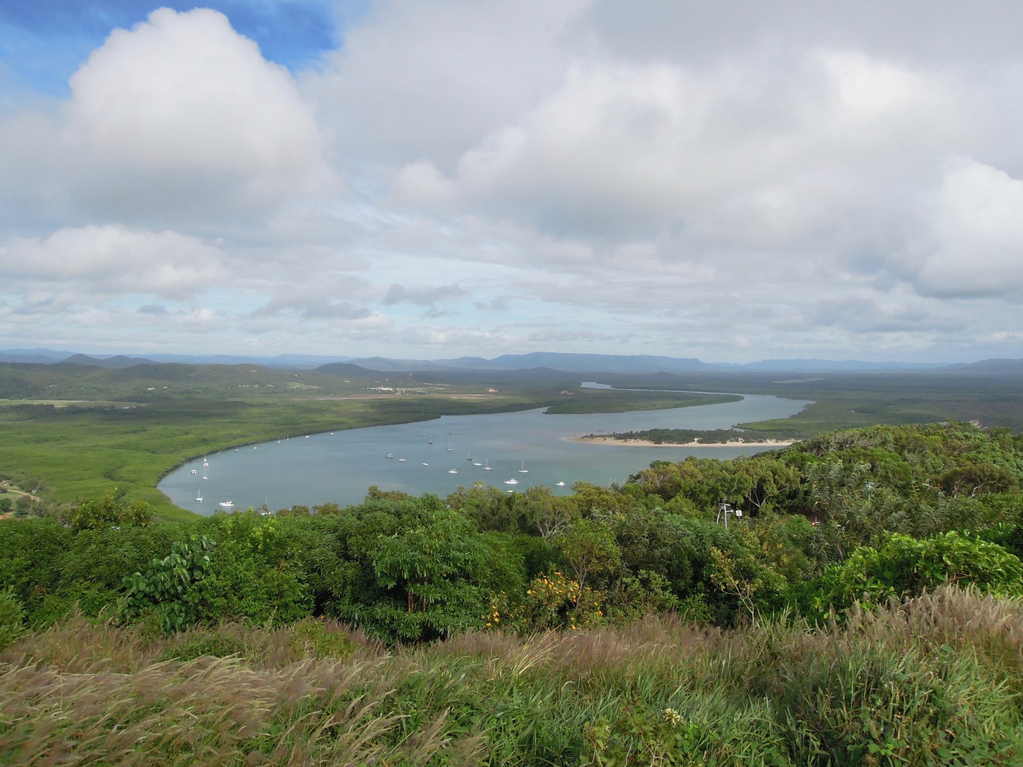 A sweeping aerial panorama of a pristine bay dotted with sailboats, surrounded by lush green vegetation and distant mountains under a partly cloudy sky. The image captures the natural beauty of tropical Australia with verdant foreground vegetation framing the tranquil anchorage below.