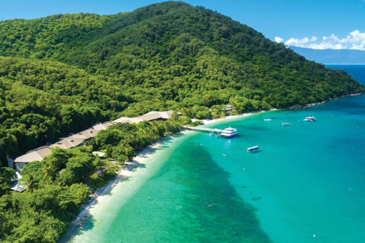An aerial view of Fitzroy Island featuring pristine turquoise waters, a sandy beach, and lush green rainforest-covered hills. Several boats are anchored in the crystal-clear bay, with a small resort or facility visible along the shoreline.