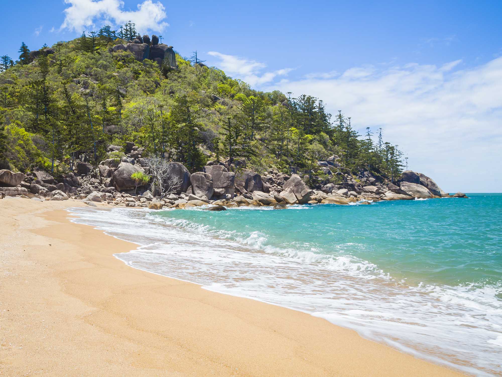 A pristine tropical beach featuring golden sand, crystal-clear turquoise waters with gentle waves, and a dramatic rocky headland covered in lush green vegetation and scattered trees. The scenic landscape includes large boulders along the shoreline and power lines visible on the hilltop, characteristic of a popular island destination.