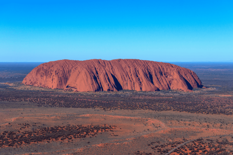 An aerial view of Uluru (Ayers Rock), the iconic red sandstone monolith in central Australia's Northern Territory, surrounded by sparse desert vegetation and spinifex grassland under a clear blue sky. The distinctive vertical striations and massive scale of this sacred Aboriginal landmark are prominently displayed against the arid landscape.