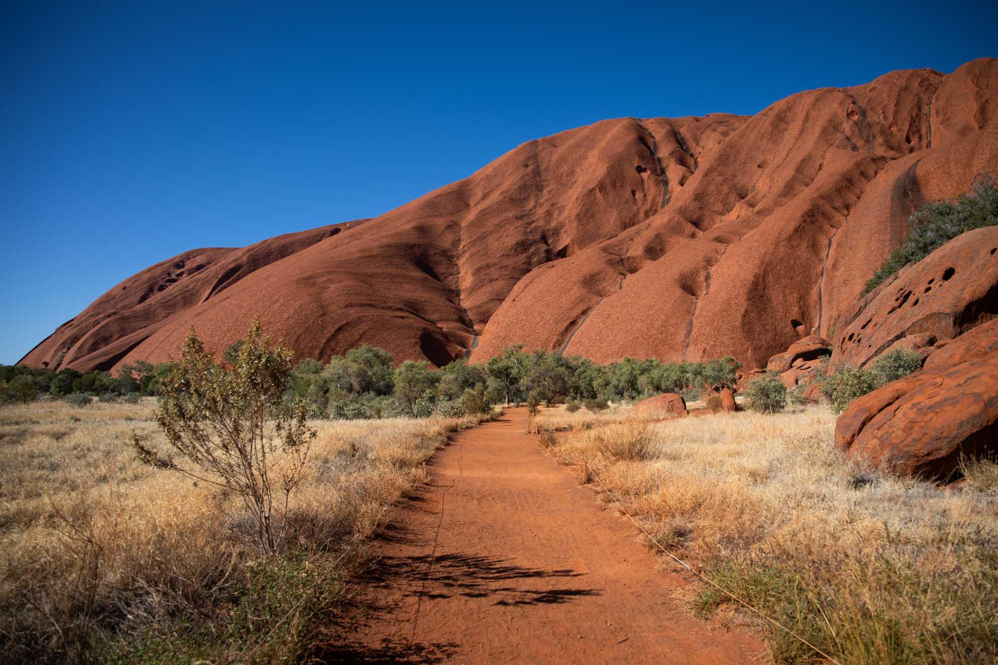 A scenic hiking trail winds through the base of Uluru (Ayers Rock) in central Australia, featuring distinctive red sandstone cliffs with natural cave formations, sparse desert vegetation, and a clear blue sky. The trail showcases the dramatic geological landscape and arid environment characteristic of Australia's iconic outback destination.