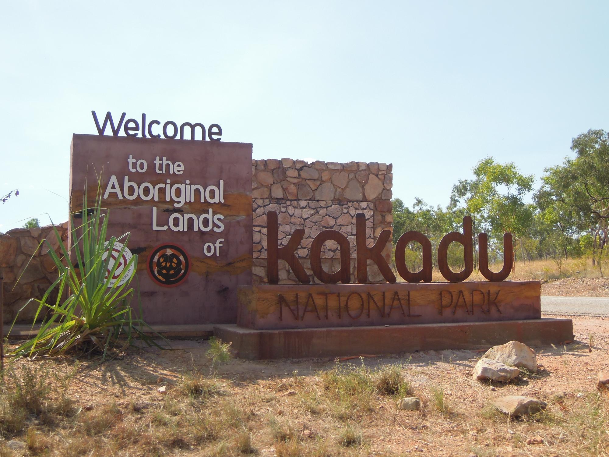 A welcoming entrance sign reading 'Welcome to the Aboriginal Lands of Kakadu National Park' constructed from stone and metal, situated in the arid Australian outback landscape with scattered vegetation and eucalyptus trees in the background.