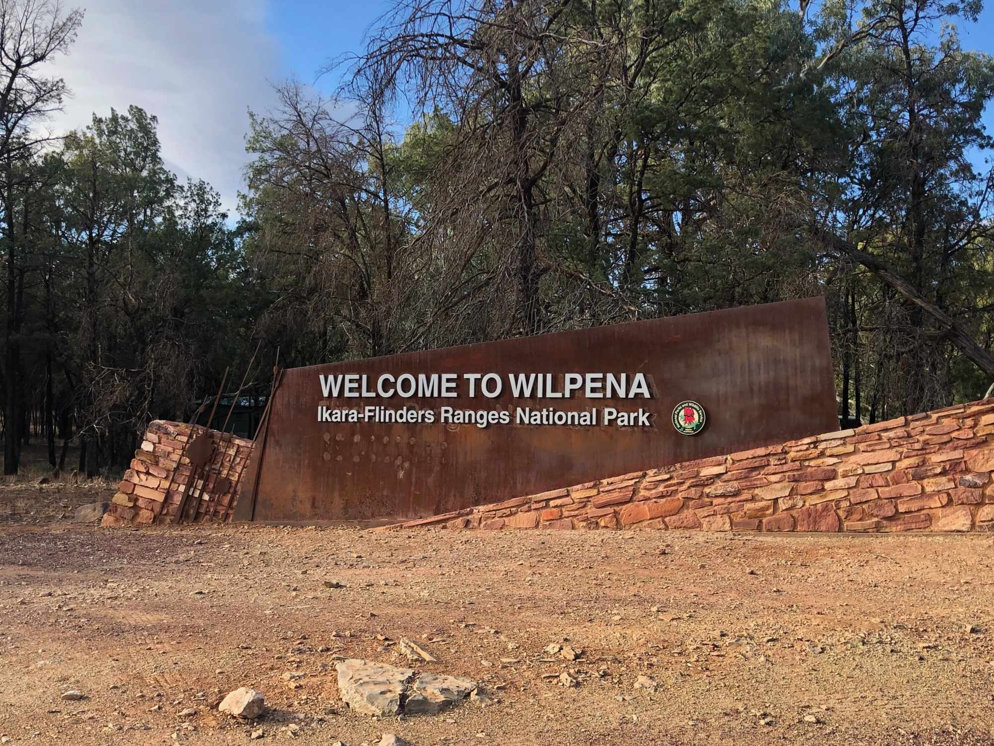 Welcome to Wilpena, Ikara-Flinders Ranges National Park A welcoming entrance sign to Wilpena in the Ikara-Flinders Ranges National Park, featuring white text on a rusted metal panel mounted on a stone base, set against a backdrop of native Australian eucalyptus trees in various stages of health.