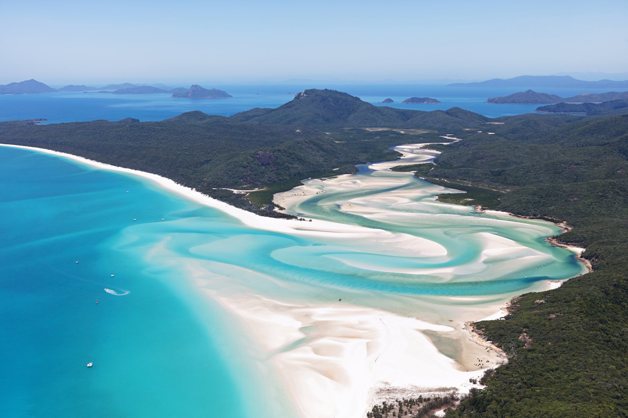 An aerial panorama of Whitehaven Beach on Whitsunday Island, Australia, showcasing the iconic white silica sand beach with turquoise lagoons, surrounding rainforest-covered hills, and numerous small islands dotting the sapphire waters of the Coral Sea.