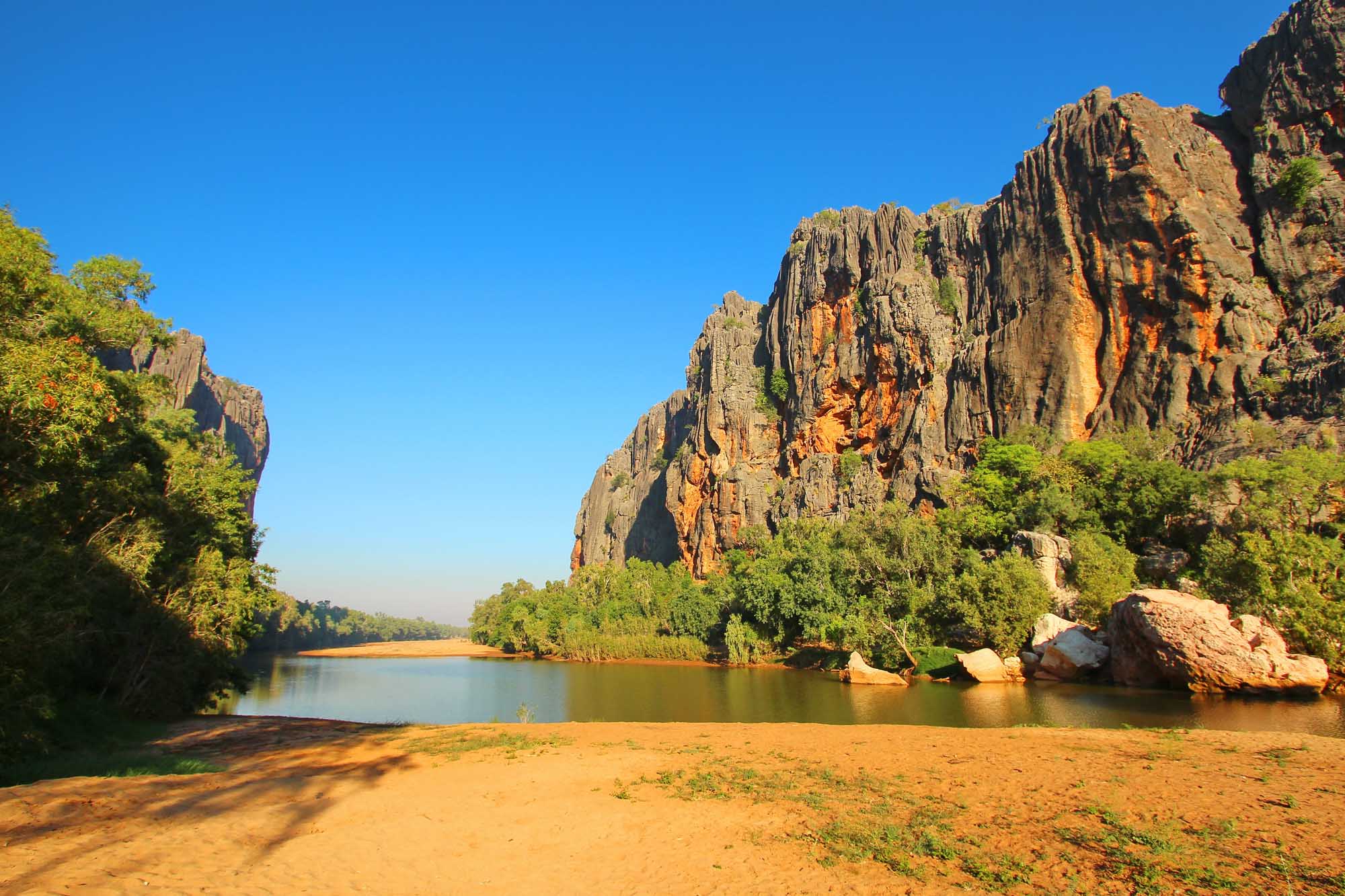 A stunning landscape of Windjana Gorge in Western Australia's Kimberley region, featuring towering columnar basalt cliffs reflected in calm river waters, with lush green vegetation framing the golden sandy beach in the foreground under a clear blue sky.