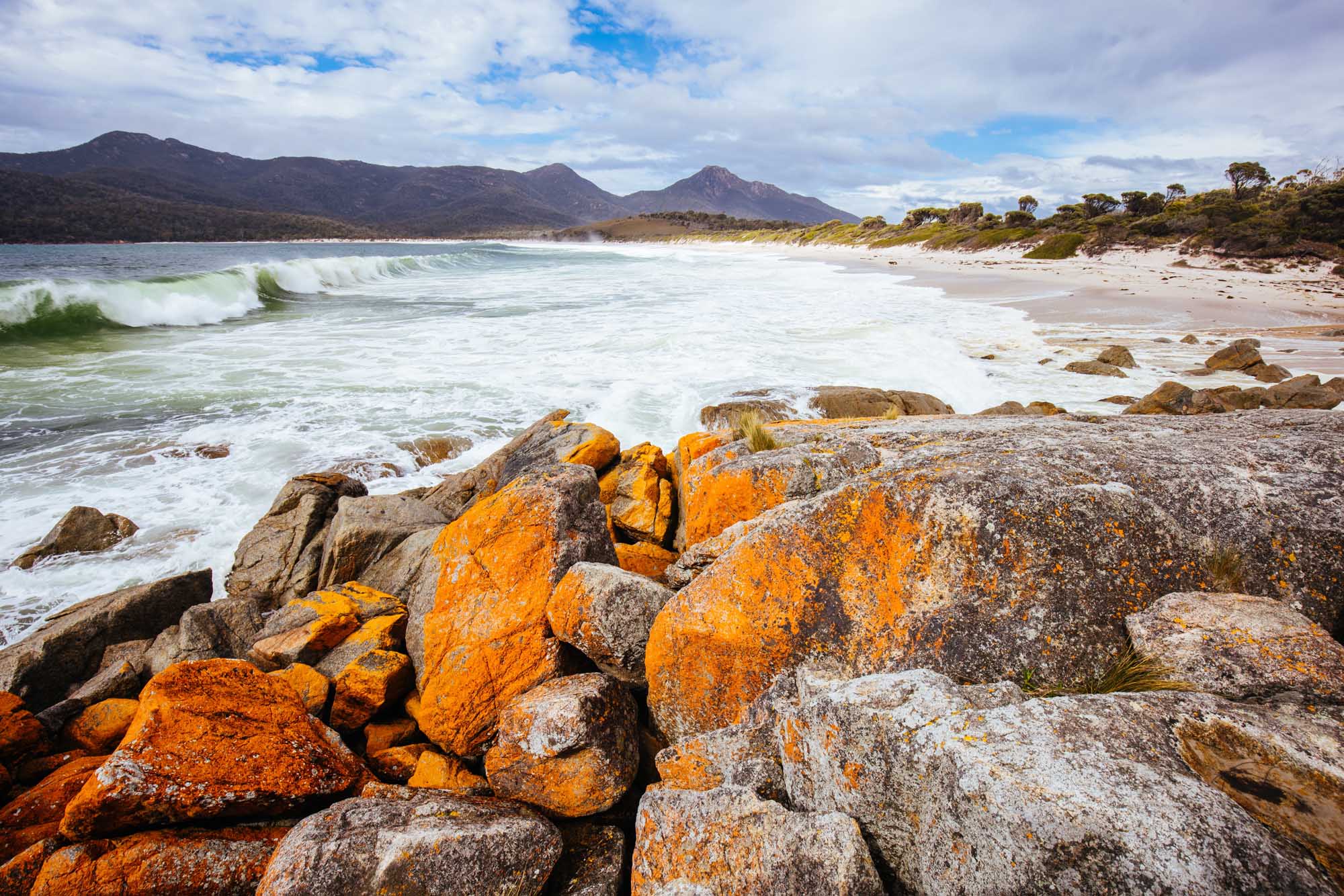 A stunning coastal landscape featuring distinctive orange lichen-covered granite boulders in the foreground of Wineglass Bay, with dramatic mountains rising behind a pristine sandy beach and rolling ocean waves under a partly cloudy sky.