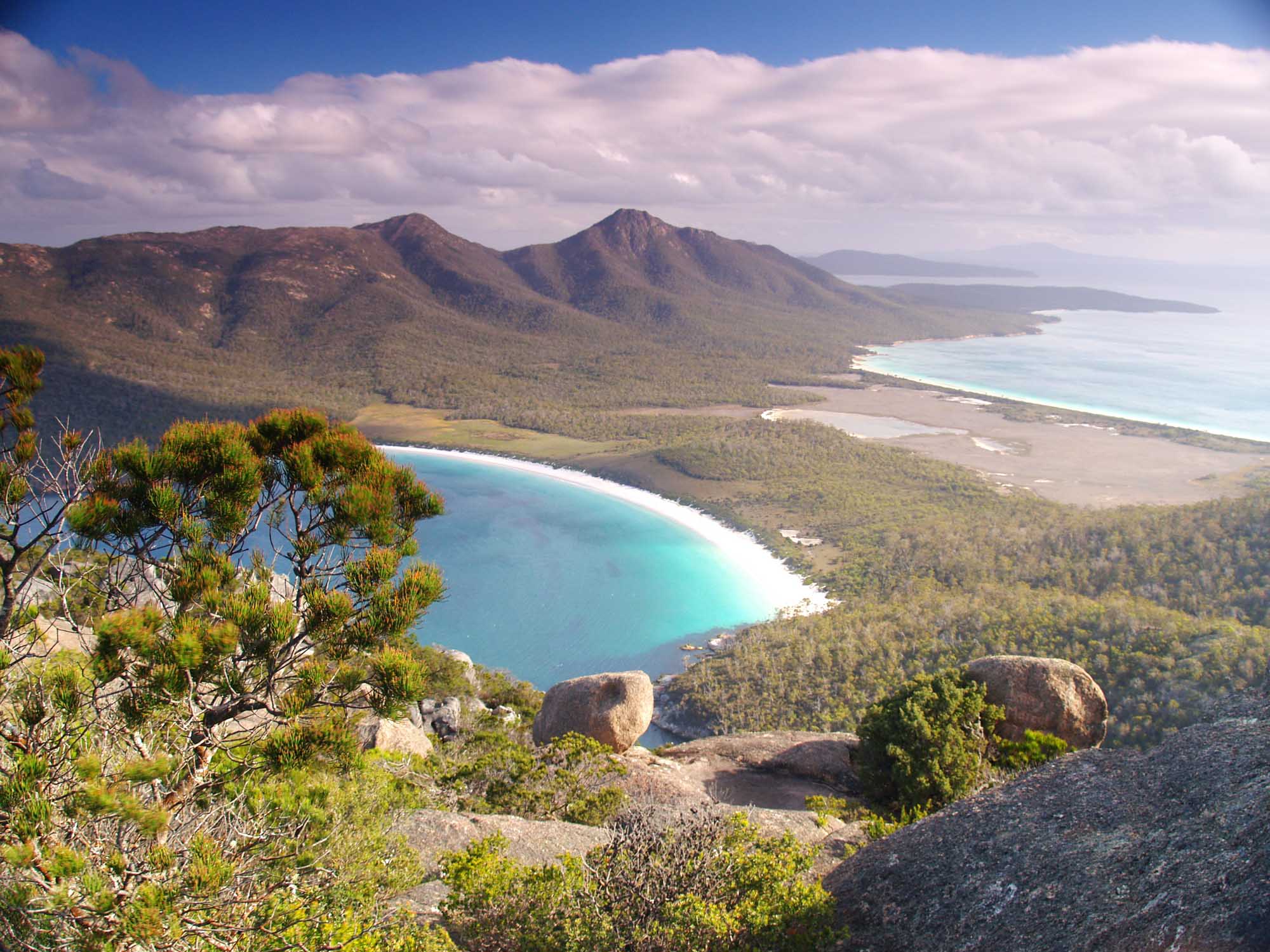 A stunning aerial view of Freycinet National Park in Tasmania, showcasing the dramatic granite peaks of the Hazards mountains, pristine turquoise waters of Wineglass Bay, and lush eucalyptus forest covering the peninsula, with dramatic cloud formations against a clear blue sky.