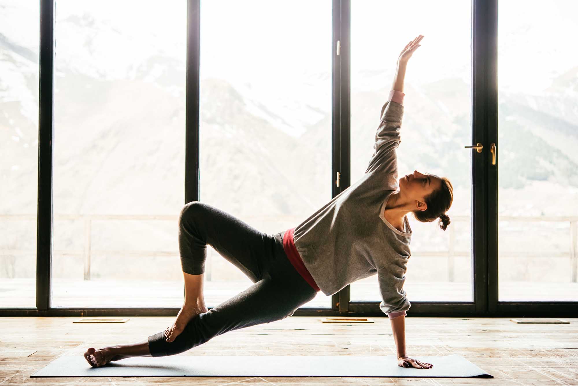 A woman performs a side plank yoga pose on a mat in front of large floor-to-ceiling windows with