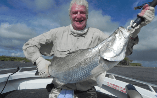 A smiling angler in light-colored fishing attire holds up a large, silvery tarpon fish on the deck of a fishing boat under a partly cloudy sky. The scene captures a successful deep-sea or saltwater fishing expedition with the angler proudly displaying his substantial catch.