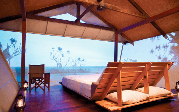 A safari-style open-air tented bedroom with a bed, wooden furniture, and lanterns, overlooking a wetland or coastal landscape at dusk.