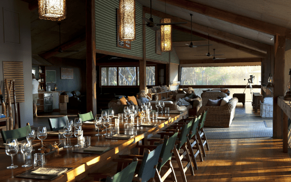 An open-air lodge dining and lounge area with a long table set for a meal, hanging lantern-style lights, and lounge seating facing large windows overlooking bushland.