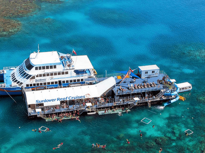 An aerial view of the Sunlover Reef Cruises ship docked at a pontoon in the crystal-clear turquoise waters of the Great Barrier Reef, with multiple passengers snorkeling and swimming in the surrounding coral reef area.