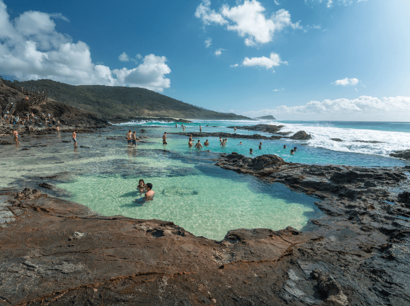 A scenic natural rock pool filled with crystal-clear turquoise water where numerous swimmers enjoy the sheltered swimming area, surrounded by dark volcanic rocks and dramatic coastal cliffs under a bright blue sky with fluffy white clouds.
