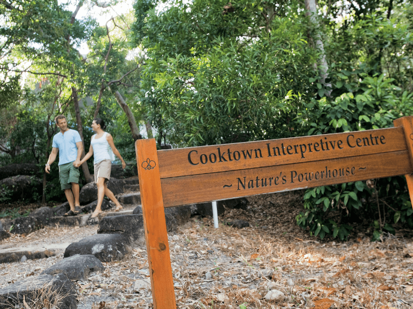 Two visitors walk along a shaded forest path at the Cooktown Interpretive Centre, passing a wooden directional sign that reads 'Cooktown Interpretive Centre ~ Nature's Powerhouse'. The lush green canopy and natural woodland setting showcase the interpretive center's focus on the local natural environment.