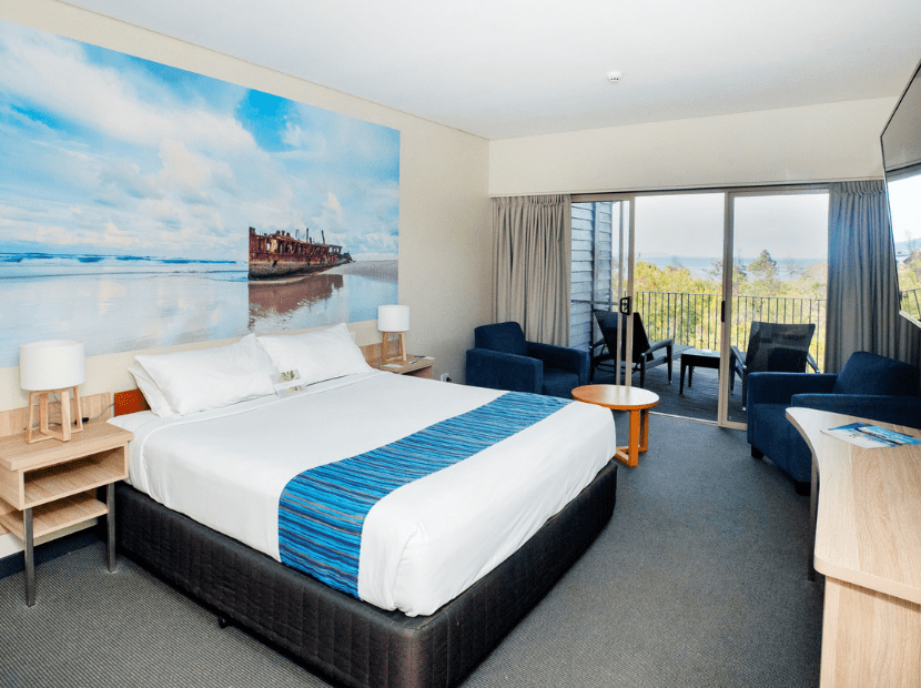 A contemporary hotel bedroom featuring a queen bed with blue accent runner, large windows opening to a scenic coastal landscape with distant mountains, and modern furnishings including comfortable seating areas. The room displays nautical-themed decor with a striking shipwreck photograph above the bed, creating a connection to maritime heritage and natural beauty.