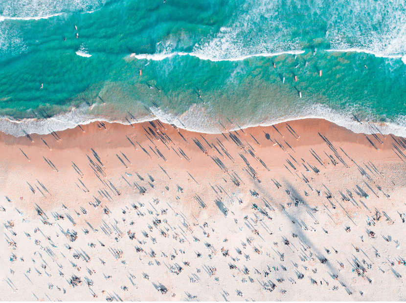 An aerial view of a pristine sandy beach populated with numerous beach umbrellas and swimmers enjoying the turquoise waters. The image captures the contrast between the pale sand, turquoise shallow waters, and deeper blue-green ocean with white wave breaks.