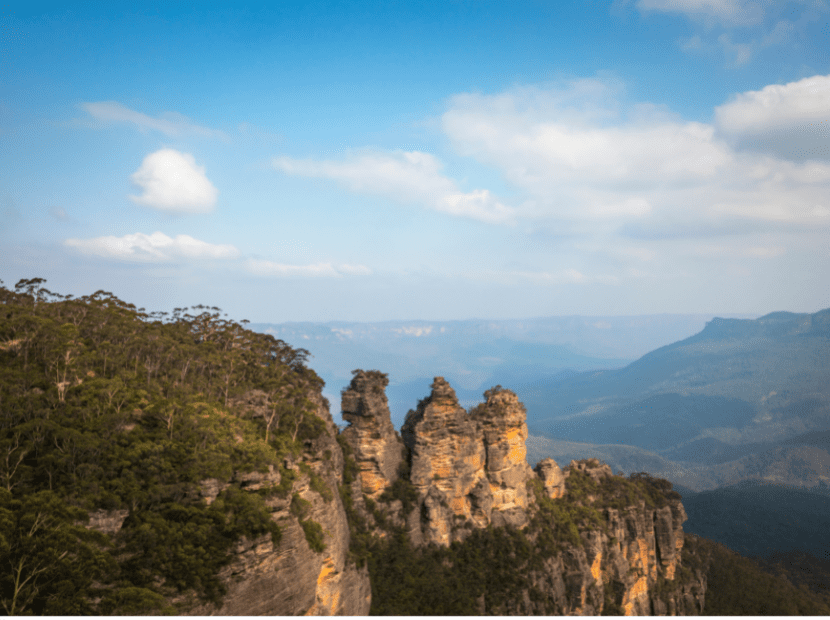 A dramatic landscape featuring three distinctive golden-hued rock pinnacles rising prominently from a forested valley, with layered blue mountains visible in the distant background under a partly cloudy sky. This iconic natural formation is surrounded by eucalyptus forest in the Blue Mountains region.