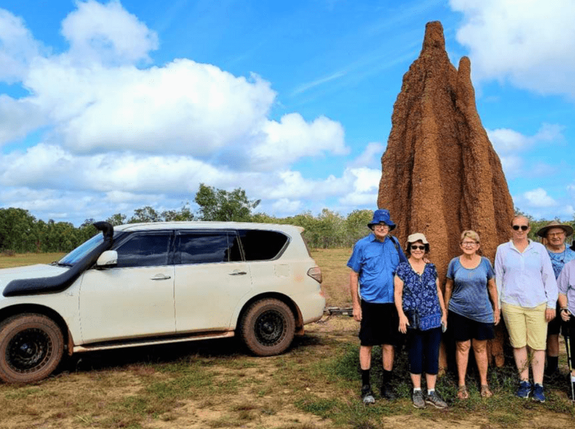 A group of six tourists pose for a photograph next to a distinctive tall termite mound in northern Australia's tropical savanna landscape. The group stands beside a white four-wheel-drive vehicle, with the iconic cone-shaped mound towering behind them against a blue sky with scattered clouds.