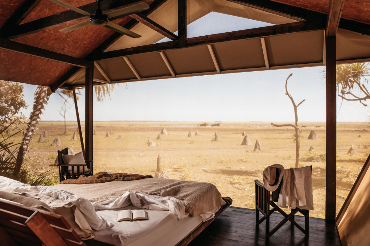 An open-air glamping tent interior with a bed and chairs looking out over a dry grassy plain dotted with termite mounds.