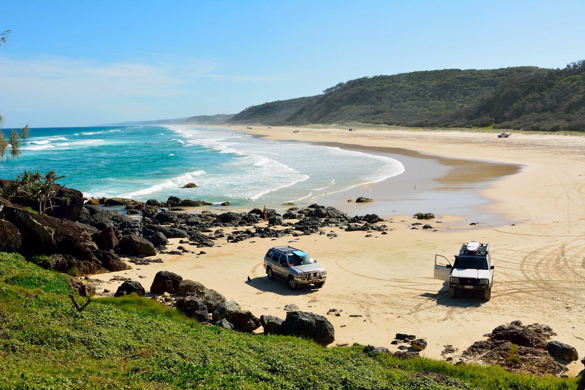 A scenic beach landscape in Great Sandy National Park, Queensland, Australia, featuring golden sand, turquoise waters, and forested hills. Two 4WD vehicles (Nissan and Toyota) are parked on the 40-mile beach with a few beachgoers visible, showcasing the popular off-road recreational destination.